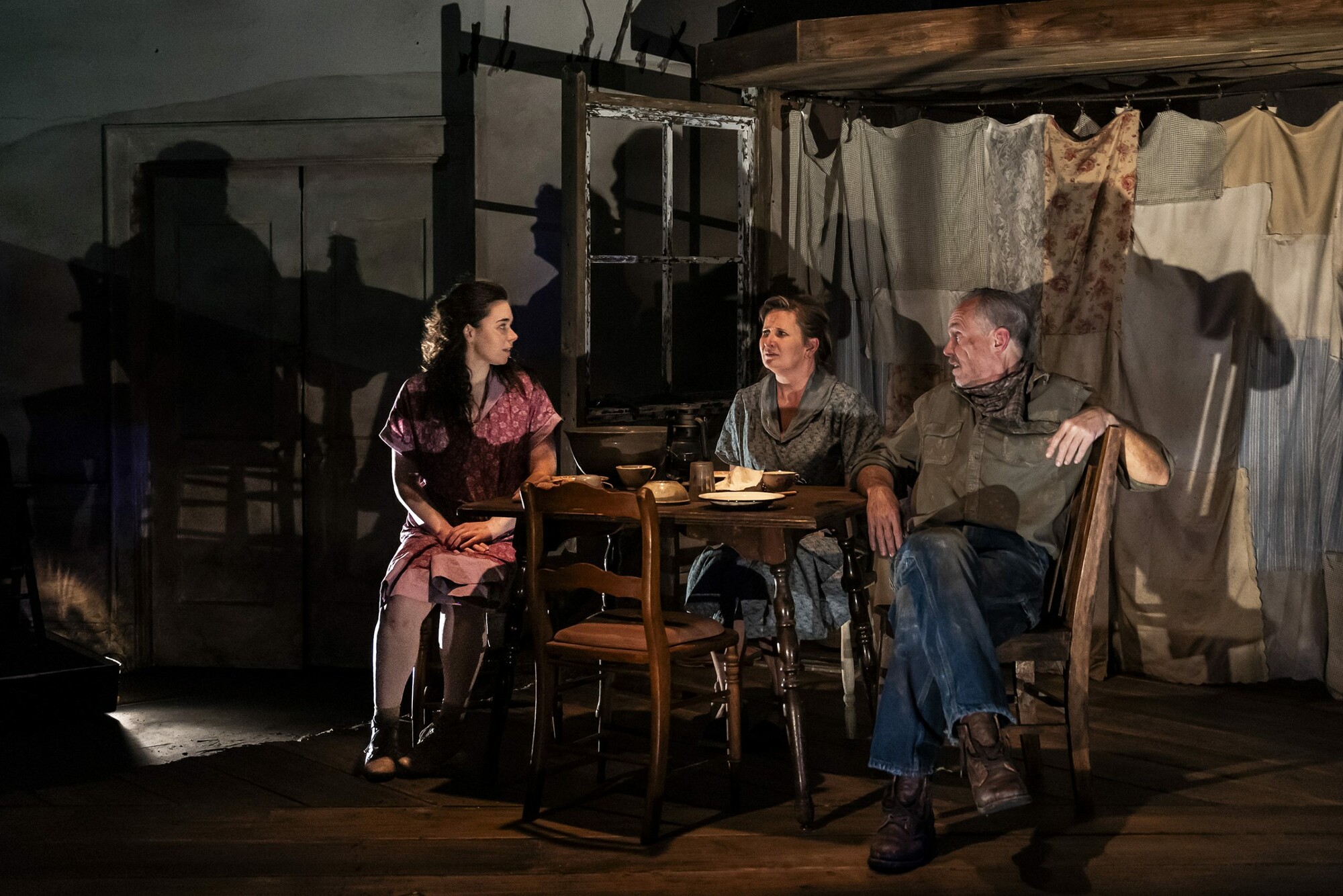 Actors sit around a table on a dimly lit set.