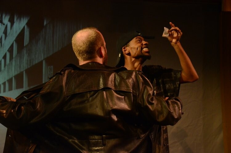 Two actor wearing black actors perform on stage in front of a projection of a concrete backdrop.