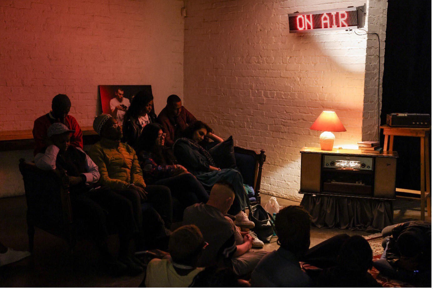 Audience members gather around a radio in a warmly lit space.