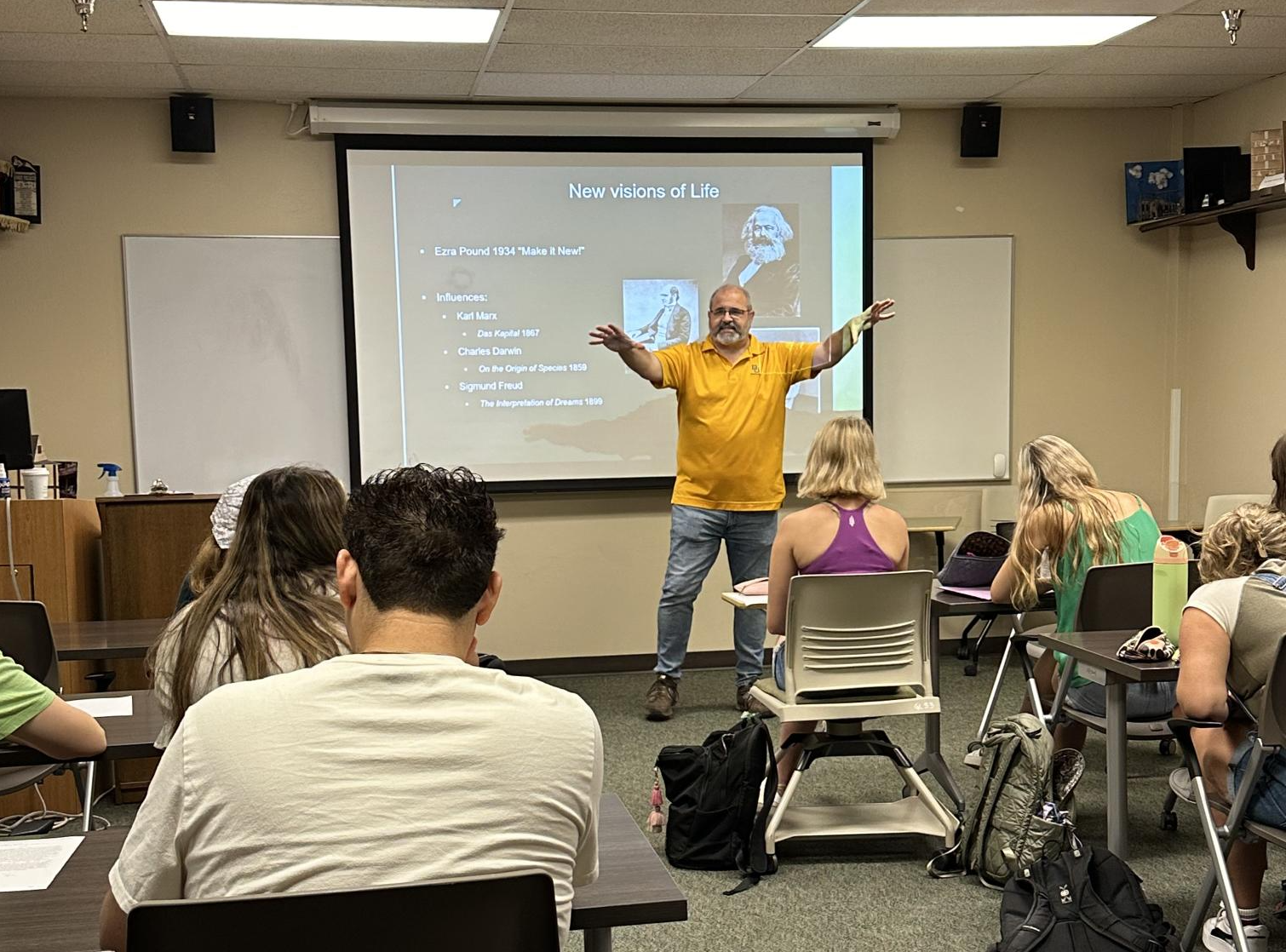 A man in an orange shirt speaks to a room full of students.