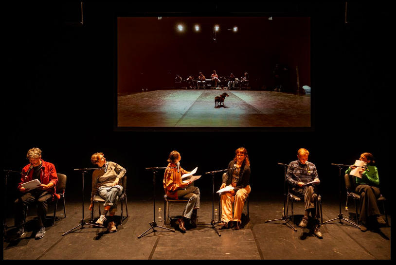 Actors perform onstage in chairs in front of a screen.