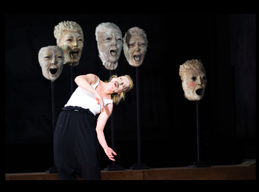 A dancer performs on stage in front of suspended grey masks.