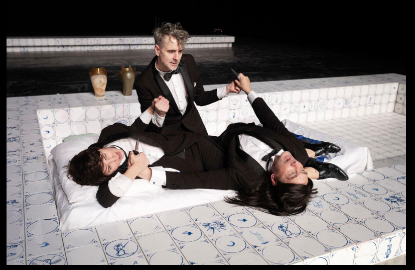 Three actors wearing suits perform on a white stage.